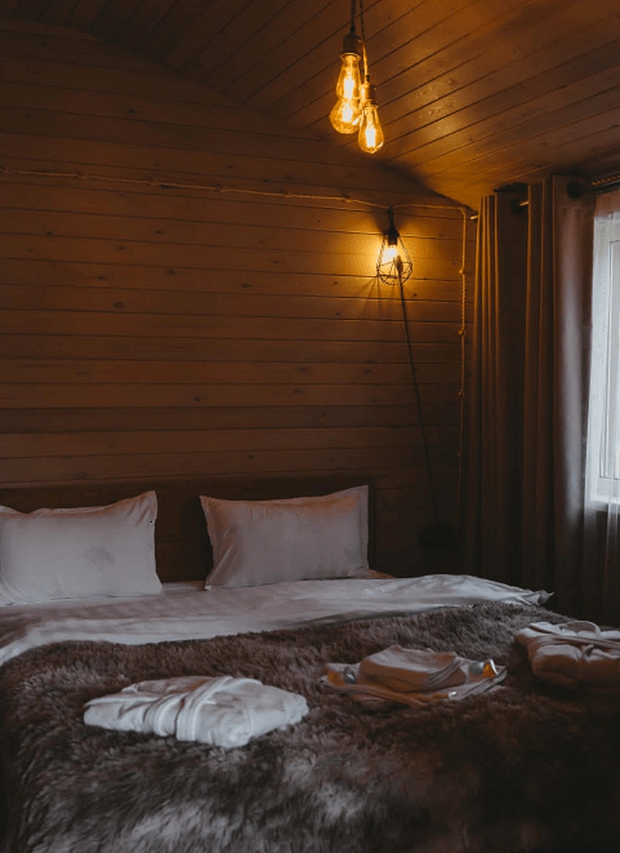 Cozy wooden hotel room with a fur-covered bed and folded bathrobes, warmly lit by hanging bulb lights
